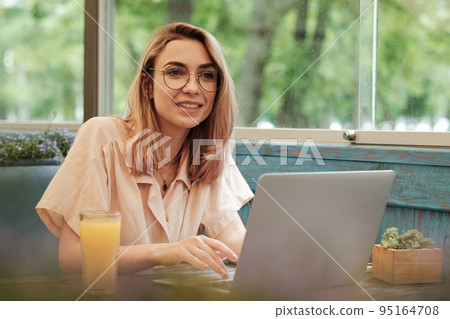 Cheerful Elegant Young Woman Working on Laptop While Sitting at a Cafe Table, Blonde Girl with Glasses, Freelancer at Work in Restaurant. 95164708