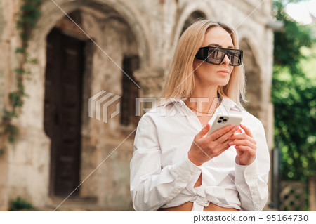 Confident Woman Texting on Smartphone While Standing on the Street, Elegant Businesswoman Wearing Sunglasses Using Mobile Phone and Looking Away. Confident Woman Texting on Smartphone While Standing on the Street, Elegant Businesswoman Wearing Sunglasses Using Mobile Phone and Looking Away. 95164730
