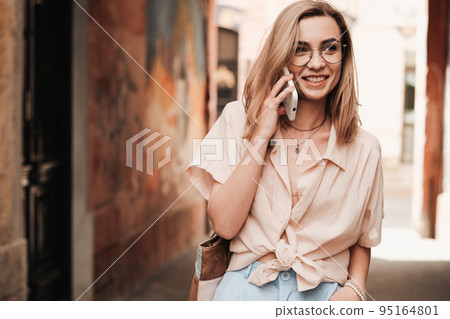 Cheerful Elegant Woman in Glasses Using Smartphone and Smiling, Stylish Blonde Girl with Eyeglasses Standing Outdoors on the Street Having Conversation Through Mobile Phone. Cheerful Elegant Woman in Glasses Using Smartphone and Smiling, Stylish Blonde Girl with Eyeglasses Standing Outdoors on the Street Having Conversation Through Mobile Phone. 95164801