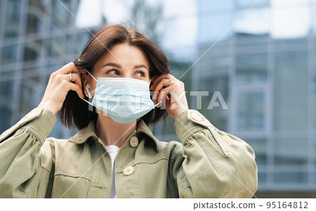 Young Woman Putting a Protective Face Mask on Her Face Because of Pandemic, Portrait of Brunette Girl Using Earphones Standing Outdoors Against City Buildings. Young Woman Putting a Protective Face Mask on Her Face Because of Pandemic, Portrait of Brunette Girl Using Earphones Standing Outdoors Against City Buildings. 95164812