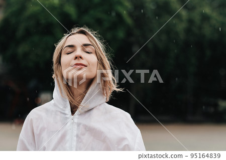 Close Up Portrait of Young Woman Wearing Raincoat While Standing on the Street at Rainy Day, Blonde Girl Dressed in Waterproof Cloak Having Fun Under Rain in City. Close Up Portrait of Young Woman Wearing Raincoat While Standing on the Street at Rainy Day, Blonde Girl Dressed in Waterproof Cloak Having Fun Under Rain in City. 95164839