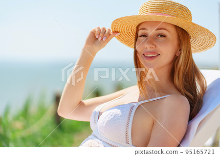 a beautiful woman in a straw hat sunbathing on a chaise longue on a beach. a beautiful woman in a straw hat sunbathing on a chaise longue on a beach. 95165721
