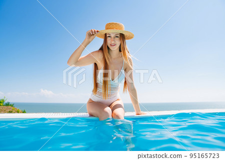 a slender woman in a white swimsuit in a straw hat sits on the pool. 95165723