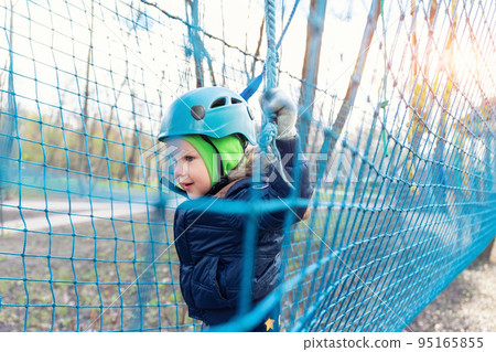 Portrait happy brave courage little toddler child boy wear safety equipment helmet enjoy passing obstacle course forest rope adventure park on cold winter day. Active outside leisure amusement camp Portrait happy brave courage little toddler child boy wear safety equipment helmet enjoy passing obstacle course forest rope adventure park on cold winter day. Active outside leisure amusement camp 95165855
