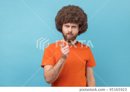 Portrait of serious man with Afro hairstyle wearing orange T-shirt standing with admonishing gesture, scolding for mistake and warning. Indoor studio shot isolated on blue background. Portrait of serious man with Afro hairstyle wearing orange T-shirt standing with admonishing gesture, scolding for mistake and warning. Indoor studio shot isolated on blue background. 95165923