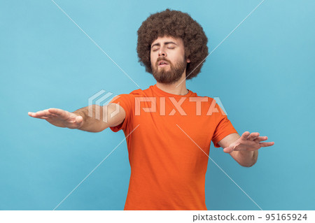 Portrait of man with Afro hairstyle moving with closed eyes, touching space with hands, confused with darkness, vision problems, eyesight disorder. Indoor studio shot isolated on blue background. Portrait of man with Afro hairstyle moving with closed eyes, touching space with hands, confused with darkness, vision problems, eyesight disorder. Indoor studio shot isolated on blue background. 95165924