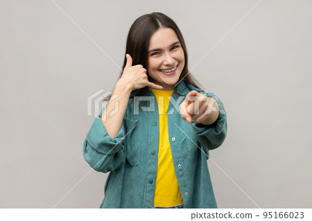 Hey you, call me back, let's contact by phone. Portrait of playful happy adult woman making telephone gesture and pointing to camera. Indoor studio shot isolated on gray background. Hey you, call me back, let's contact by phone. Portrait of playful happy adult woman making telephone gesture and pointing to camera. Indoor studio shot isolated on gray background. 95166023