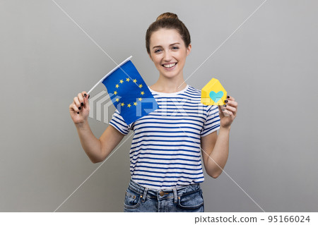 EU immigration. Portrait of delighted woman wearing striped T-shirt holding flag of European Union, travel and rental housing in Europe. Indoor studio shot isolated on gray background. EU immigration. Portrait of delighted woman wearing striped T-shirt holding flag of European Union, travel and rental housing in Europe. Indoor studio shot isolated on gray background. 95166024