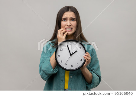 Portrait of impatient nervous woman with dark hair biting her nails and holding big wall clock, deadline, wearing casual style jacket. Indoor studio shot isolated on gray background. Portrait of impatient nervous woman with dark hair biting her nails and holding big wall clock, deadline, wearing casual style jacket. Indoor studio shot isolated on gray background. 95166040