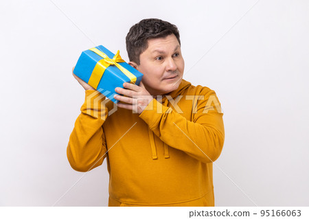 Portrait of curious middle aged man holding and shaking wrapped present box, being interested what inside, wearing urban style hoodie. Indoor studio shot isolated on white background. 95166063