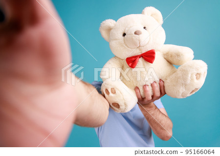 Portrait of unknown bearded man making selfie or broadcasting livestream, point of view photo, hiding his face behind soft teddy bear. Indoor studio shot isolated on blue background. 95166064