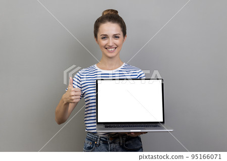Happy smiling satisfied woman wearing striped T-shirt holding laptop with empty blank display and showing thumbs up, pleased with online application. Indoor studio shot isolated on gray background. Happy smiling satisfied woman wearing striped T-shirt holding laptop with empty blank display and showing thumbs up, pleased with online application. Indoor studio shot isolated on gray background. 95166071