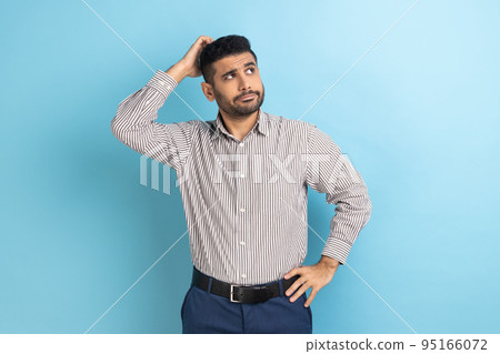 Portrait of young pensive businessman with beard touching head and thinking about important question, making hard decision, wearing striped shirt. Indoor studio shot isolated on blue background. Portrait of young pensive businessman with beard touching head and thinking about important question, making hard decision, wearing striped shirt. Indoor studio shot isolated on blue background. 95166072