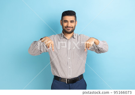 Cheerful satisfied businessman pointing fingers down, paying attention to advertising area, blogger recommending to subscribe, wearing striped shirt. Indoor studio shot isolated on blue background. Cheerful satisfied businessman pointing fingers down, paying attention to advertising area, blogger recommending to subscribe, wearing striped shirt. Indoor studio shot isolated on blue background. 95166073