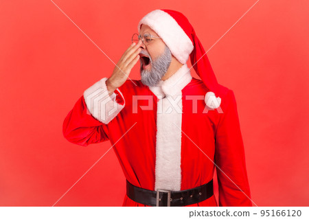 Elderly man with gray beard wearing santa claus costume yawning and covering mouth with arm, feeling drowsy after sleepless night, lack of energy. Indoor studio shot isolated on red background. 95166120