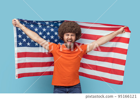 Portrait of man with Afro hairstyle in T-shirt holding USA flag and looking at camera with rejoicing look, screaming, celebrating national holiday. Indoor studio shot isolated on blue background. 95166121
