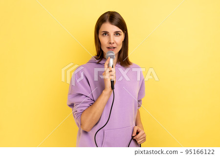 Young adult beautiful woman reporter holding microphone, reporting and telling opinion, discussing important topics, wearing purple hoodie. Indoor studio shot isolated on yellow background. 95166122