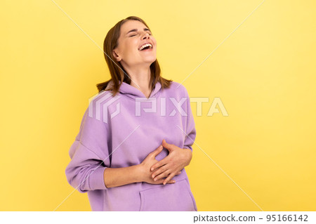 Portrait of happy woman with dark hair holding belly, laughing, hearing funny joke or anecdote, being in good mood, wearing purple hoodie. Indoor studio shot isolated on yellow background. Portrait of happy woman with dark hair holding belly, laughing, hearing funny joke or anecdote, being in good mood, wearing purple hoodie. Indoor studio shot isolated on yellow background. 95166142