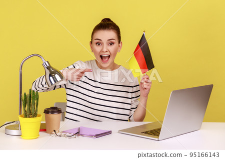 Portrait of young adult attractive woman office employee sitting at workplace and pointing at German flag, job or education abroad. Indoor studio studio shot isolated on yellow background. 95166143