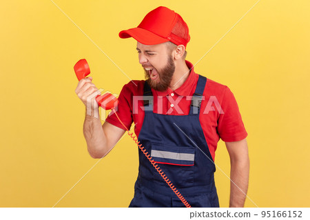 Portrait of angry aggressive worker wearing blue overalls screaming to client on handset, does not accept order, incompetent service personnel. Indoor studio shot isolated on yellow background. 95166152
