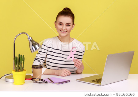Optimistic pretty woman volunteer sitting at workplace with laptop and showing pink ribbon, symbol of breast cancer awareness, female health. Indoor studio studio shot isolated on yellow background. Optimistic pretty woman volunteer sitting at workplace with laptop and showing pink ribbon, symbol of breast cancer awareness, female health. Indoor studio studio shot isolated on yellow background. 95166166