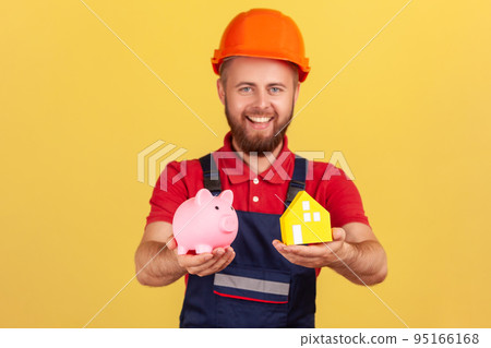 Portrait of satisfied builder man wearing blue uniform and protective helmet showing piggy bank and paper house, looking at camera. Indoor studio shot isolated on yellow background. Portrait of satisfied builder man wearing blue uniform and protective helmet showing piggy bank and paper house, looking at camera. Indoor studio shot isolated on yellow background. 95166168