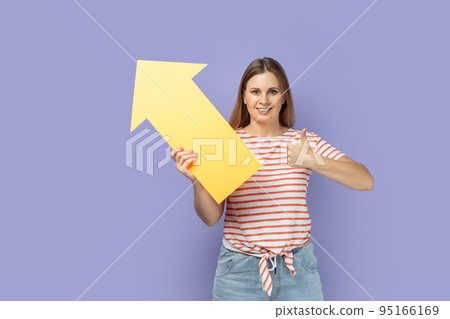 Portrait of pretty smiling blond woman wearing striped T-shirt holding big yellow arrow pointing aside and showing thumb up, like gesture. Indoor studio shot isolated on purple background. 95166169