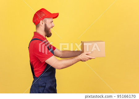 Profile portrait of positive deliveryman holding cardboard parcel, delivering order door-to-door, shipment and cargo transportation service. Indoor studio shot isolated on yellow background. Profile portrait of positive deliveryman holding cardboard parcel, delivering order door-to-door, shipment and cargo transportation service. Indoor studio shot isolated on yellow background. 95166170