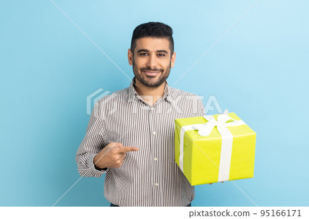 Happy businessman with beard standing, holding yellow gift box, pointing finger, looking at camera with toothy smile, wearing striped shirt. Indoor studio shot isolated on blue background. Happy businessman with beard standing, holding yellow gift box, pointing finger, looking at camera with toothy smile, wearing striped shirt. Indoor studio shot isolated on blue background. 95166171