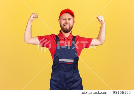 Portrait of proud strong worker man wearing blue uniform and red cap standing with raised arms and showing his power, looking at camera with pride. Indoor studio shot isolated on yellow background. 95166193