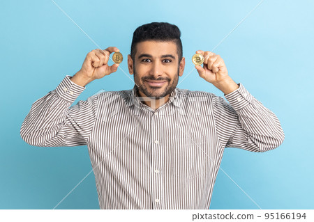 Cryptocurrency btc coin. Rich satisfied happy businessman looking at camera, holds golden bitcoin, electronic money, wearing striped shirt. Indoor studio shot isolated on blue background. 95166194