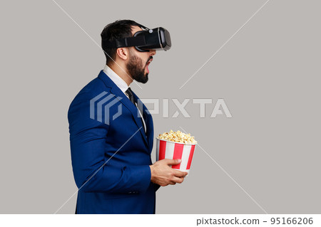 Side view of excited amazed bearded businessman in VR headset watching movie with popcorn, keeps mouth open, wearing official style suit. Indoor studio shot isolated on gray background. Side view of excited amazed bearded businessman in VR headset watching movie with popcorn, keeps mouth open, wearing official style suit. Indoor studio shot isolated on gray background. 95166206