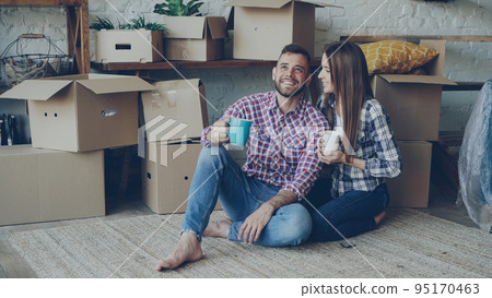 Girlfriend and boyfriend are talking making plans sitting on floor of new purchased apartment, looking around and holding mugs. Relationship, people and relocation concept. Girlfriend and boyfriend are talking making plans sitting on floor of new purchased apartment, looking around and holding mugs. Relationship, people and relocation concept. 95170463