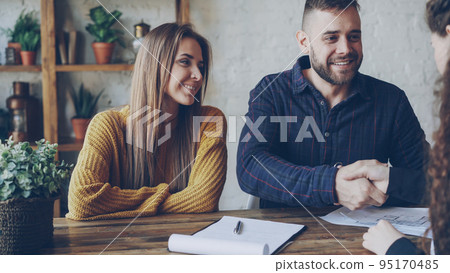 Attractive female broker is selling house to young couple, people are signing documents and shaking hands, realtor is giving key to new owner, he is kissing his wife. 95170485