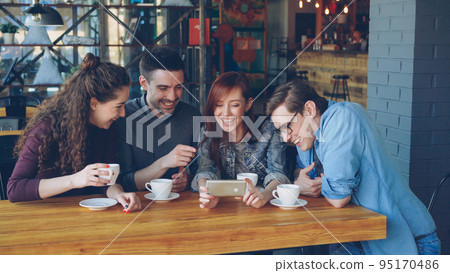 Happy good-looking students are watching funny video, discussing it and laughing during meeting in cafe. Cheerful people, modern technology and eating out concept. Happy good-looking students are watching funny video, discussing it and laughing during meeting in cafe. Cheerful people, modern technology and eating out concept. 95170486