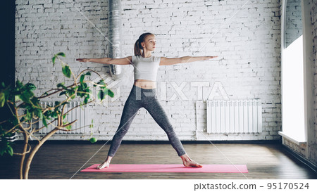 Young slim blond girl is doing yoga complex in nice studio with white walls. She is starting with warrior poses, then bending forward and backward and finishing with namaste. 95170524
