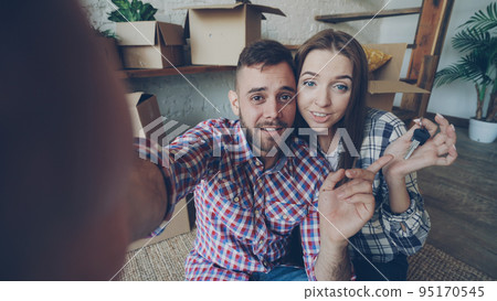 Happy young couple is making video call with smartphone after relocation. They are greeting friends, showing new house keys and many boxes, chatting and smiling. 95170545