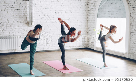 Three pretty young ladies are practicing Lord of the dance pose Natarajasana while doing yoga in modern wellness center with hign ceiling. Concentration flexibility energy concept. Three pretty young ladies are practicing Lord of the dance pose Natarajasana while doing yoga in modern wellness center with hign ceiling. Concentration flexibility energy concept. 95170620