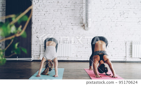 Young blond girl is enjoying individual yoga practice with friendly female instructor in light studio. Women are doing sequence of asanas on bright mats. Friendly relaxing atmosphere. 95170624