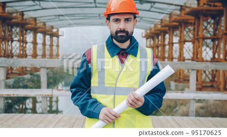 Portrait of male builder wearing helmet and vest standing outside construction site holding blueprint and looking at camera. People and profession concept. 95170625