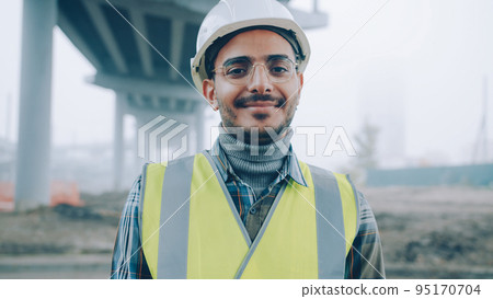 portrait of cheerful Arab man wearing safety helmet and vest smiling standing against urban construction background alone and looking at camera portrait of cheerful Arab man wearing safety helmet and vest smiling standing against urban construction background alone and looking at camera 95170704