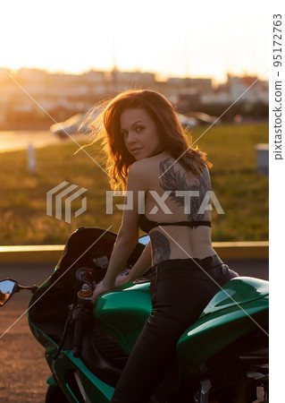 Confident young woman sitting on motorbike on parking area Confident young woman sitting on motorbike on parking area 95172763