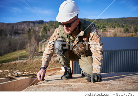 Man worker building wooden frame house. Carpenter using tape measure for measuring OSB sheet on rooftop. Carpentry concept. 95173753