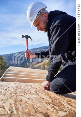 Carpenter hammering nail into OSB panel on the roof top of future cottage. Man worker building wooden frame house. Carpentry and construction concept. 95173758