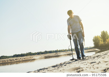 Man with metal detector walks along the sandy river bank. Search for treasures and metal for recycling Man with metal detector walks along the sandy river bank. Search for treasures and metal for recycling 95175506