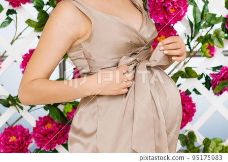 A pregnant woman ties a bow on her belly dress on a background with peony flowers on the wall. A young woman in a beige satin maternity dress is waiting for the birth of a child. Pregnancy, 95175983