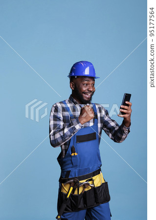 African american builder excited by news of new construction contract received by video call on cell phone. Contractor celebrating with emotion holding mobile device against blue background. 95177564