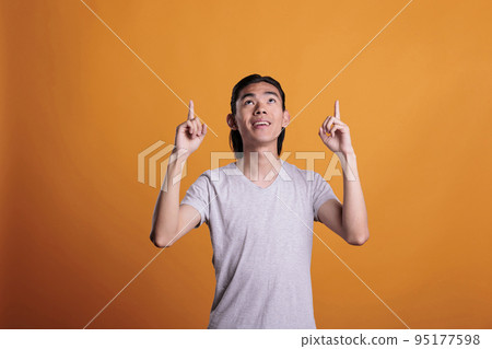 Young smiling asian man pointing up with forefingers, advertising product. Cheerful teenager standing, looking upwards, showing promotion, studio medium shot on orange background 95177598