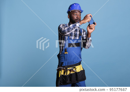 African american builder taking a break at work playing video games on a cell phone. Construction worker wearing work uniform relaxing after a busy day against blue background in studio shot. 95177687
