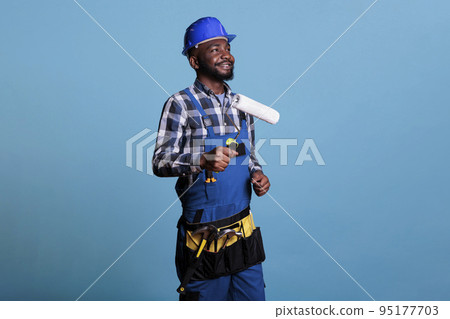 Happy construction worker holding paint roller performing interior renovation with optimism. House painter dressed in uniform and protective helmet against blue background, studio shot 95177703